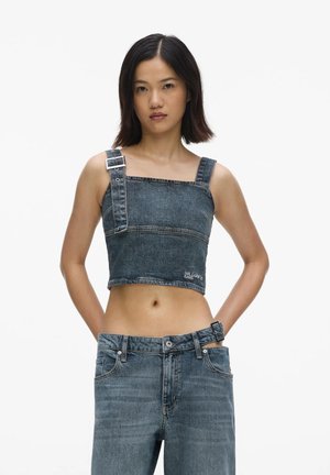 Femme portant un crop top en denim foncé assorti avec une bride à boucle et un jean taille haute avec une ceinture à boucle réglable, debout sur un fond blanc.
