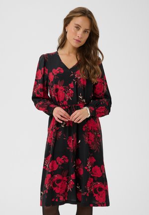Femme aux longs cheveux bruns portant une robe noire avec de grands motifs floraux rouges, debout les mains jointes devant elle.