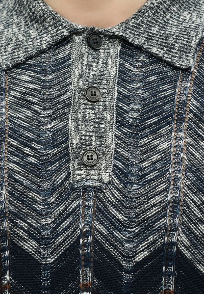 Πλεκτό πουλόβερ με γιακά σε μπλε, λευκό και γκρι σχέδιο ζιγκ-ζαγκ, με τρία μαύρα κουμπιά στη μπροστινή πατιλέτα.