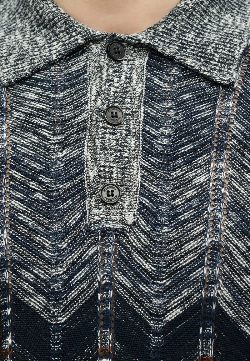 Collared knit sweater in blue, white, and gray chevron pattern with three black buttons down the front placket.