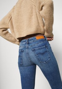Jeans de mezclilla azul con un diseño ajustado, con bolsillos traseros y una etiqueta de cuero marrón en la cintura. Suéter de punto en un beige claro.