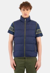 Marinblå pufferväst med gröna detaljer, dragkedjestängning och korta ärmar. Har horisontella ränder på ärmarna och sidfickor.