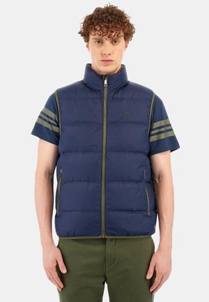 Marineblå puffer vest med grønne detaljer, lynlåslukning og korte ærmer. Har vandrette striber på ærmerne og sidelommer.