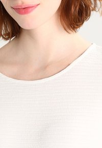White textured top with a wide neckline, featuring a ribbed pattern and soft fabric that drapes gently. Close-up view of the shoulders and neck.