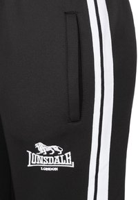 Černé joggingové kalhoty s bílými postranními pruhy, obsahující malé bílé vyšívané logo lva a dvě boční kapsy. Měkká textilie.