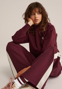 Fiatal nő hullámos hajjal, bordó melegítőnadrágban, fehér oldalsávokkal, keresztbe tett lábakkal ül, és a kezére támasztja a fejét.