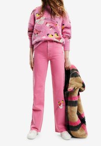 Personne portant un pantalon large rose avec un visage de la Panthère Rose, un pull assorti de la Panthère Rose, des baskets blanches, tenant un manteau en fausse fourrure à carreaux.