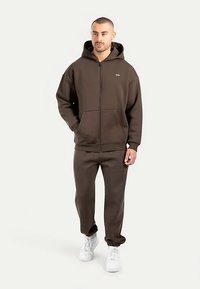 Brun fleece-hoodie med lynlås, der har en kængurulomme og ribbede ærmegab, kombineret med matchende joggingbukser og hvide sneakers.