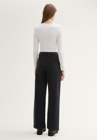 Jeans anchos negros con bolsillos traseros, que presentan una cintura alta y costuras sutiles. Combinados con una camisa blanca de manga larga.