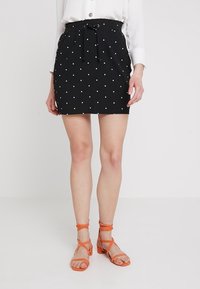 Falda negra con lunares blancos y cintura ajustable, combinada con sandalias naranjas de tiras. Tejido suave y diseño entallado.