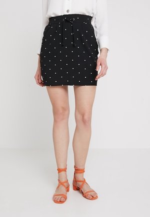 Jupe noire à pois blancs avec une taille à cordon, portée avec des sandales orange à lanières. Tissu lisse et design ajusté.