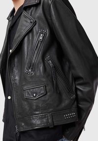 Schwarze Leder-Motorradjacke mit mehreren Reißverschluss-Taschen, genagelten Akzenten und einer leicht strukturierten Oberfläche. Mit klassischem Kragen und seitlichen Druckknöpfen.