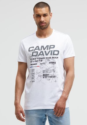Mann trägt weißes Grafik-T-Shirt mit "Camp David" und Kartendesign, kombiniert mit hellblauen Jeans, steht vor schlichtem Hintergrund.
