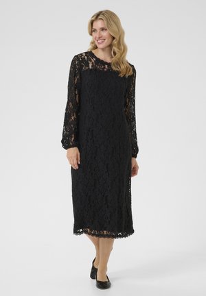 Abito midi in pizzo nero con maniche lunghe, orlo scallopato e motivo floreale. Tessuto leggero che offre un aspetto texturizzato. Indossato con ballerine nere.