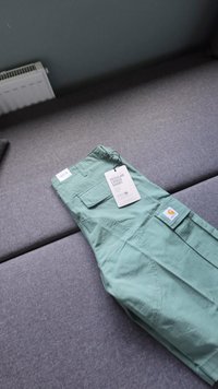 Pantaloni cargo verde chiaro in cotone, dotati di tasche, etichetta e patch con logo. Adagiati su una superficie di tessuto grigio.