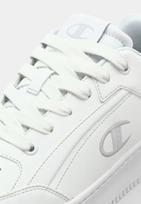 White sneaker with textured leather, flat white laces, and a gray logo on the side. Rounded toe and subtle stitching details.