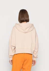 Cropped hoodie in light beige cotton with a hood, dropped shoulders, and a ribbed hem. Paired with orange sweatpants.