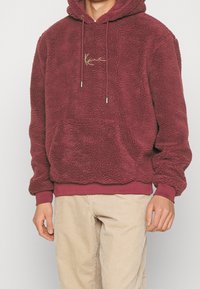 Sudadera con capucha de terciopelo burdeos con una superficie texturizada, bolsillo tipo canguro, bordado de logo dorado y capucha con cordón. Combinada con pantalones de pana beige.