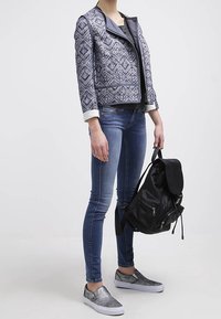 Person med mönstrad blå jacka, slimfit jeans, grå slip-on skor, som håller en svart ryggsäck vid handtaget på sidan.