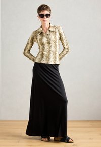 Femme aux cheveux courts et lunettes de soleil, portant une chemise à manches longues beige à motifs, une jupe maxi noire et des sandales noires, se tenant sur un sol en bois.