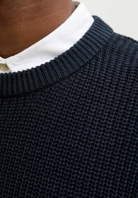 Námořnický pletený svetr s hrdlem s žebrováním, texturovanou tkaninou a bílou košili pod ním. Detailní pohled na výstřih a materiál.