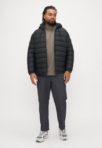 Schwarze Steppjacke mit Kapuze, graues T-Shirt mit Rundhalsausschnitt, dunkelgraue Hose mit Seitentaschen und graue Sneakers mit weißen Akzenten.