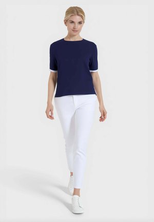 Donna bionda che cammina, indossa una maglietta blu navy a maniche corte, pantaloni bianchi aderenti e sneakers bianchi su uno sfondo chiaro e uniforme.