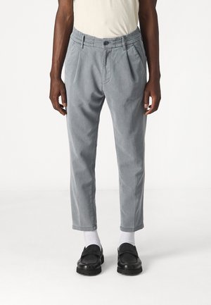 Man draagt lichtgrijze geplooide broek, witte sokken, zwarte loafers en een ingestopt wit T-shirt, staand tegen een effen witte achtergrond.