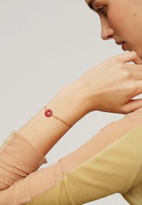 Pulsera dorada con un acento de cuenta circular rosa. La banda presenta un diseño de cadena delicada. Lleva puesta en una muñeca ligeramente bronceada.