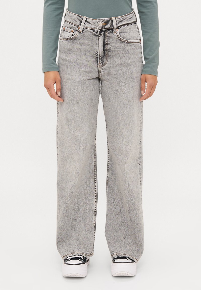 Jeans de tiro alto y pierna ancha en gris claro, con un acabado desgastado. Presentan cinco bolsillos, pasadores para cinturón y costuras en contraste. Combinados con zapatillas de plataforma.