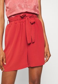 Shorts rouges taille haute avec une ceinture nouée, en tissu texturé et avec des poches latérales, portés avec un blouse rose ornée de broderie décorative.