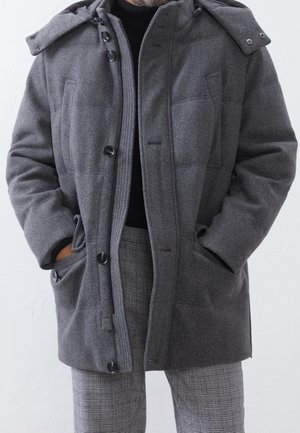 Mann trägt einen grauen Wollmantel mit großen Knöpfen über einem schwarzen Rollkragenpullover und grauen karierten Hosen, Hände in den Manteltaschen.