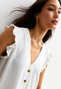 Vit blus med volangärmar, gjord av textil med struktur, med en djup V-halsringning och tre bruna knappar ner längs framsidan. Gyllene kedjehalsband synligt.