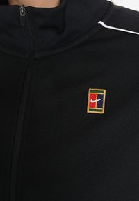 Musta vetoketjullinen urheilu takki, joka on valmistettu teksturoidusta kankaasta ja jossa on monivärinen brodeerattu Nike-logo rintamuksessa ja valkoisia yksityiskohtia.