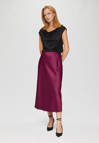Zwart gedrapeerde top gecombineerd met een satijnen bordeauxrode midi rok. De outfit heeft een ontspannen pasvorm en een slanke silhouet, met puntige zwarte hakken.