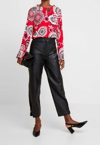 Top rojo de flores con mangas largas y puños con volantes, combinado con pantalones de cuero negros de cintura alta y tacones negros de punta, sosteniendo un bolso de mano.