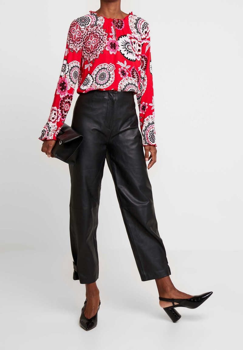 Top rojo de flores con mangas largas y puños con volantes, combinado con pantalones de cuero negros de cintura alta y tacones negros de punta, sosteniendo un bolso de mano.