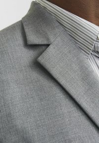 Pilkas audinio bleizeris su tekstūruotu paviršiumi, turintis aštrų apykaklę ir po juo vilkintis raštuotas dryžuotas marškinys.