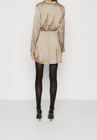 Vestido de camisa de satén beige con mangas largas, cintura elástica y una textura suave, combinado con medias opacas negras y tacones.