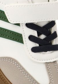 Zapatilla blanca con rayas verdes y suela gris, con cordones negros y una correa de velcro. Fabricada con material sintético suave.