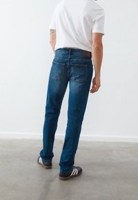 Blue denim jeans with a straight fit, five-pocket design, and brown leather patch at the back waistband, paired with black sneakers.