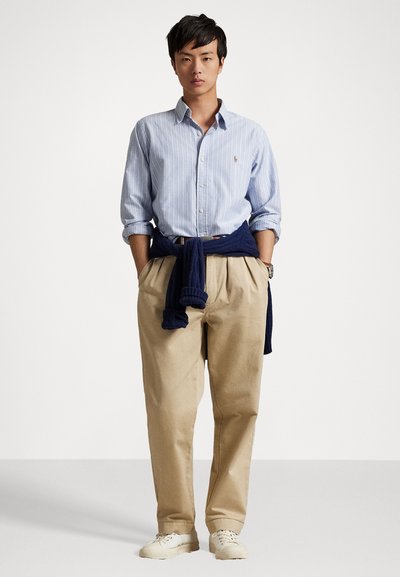 Lichtblauw gestreepent overhemd met knopen en een klein logo, beige broek, en een marineblauwe trui om de taille geknoopt. Witte sneakers maken de outfit af.