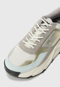 Sneaker aus einer Materialkombination, einschließlich grauem Wildleder, Mesh und metallischen Akzenten. Die Farbpalette umfasst Weiß, Grau, Hellblau und Schwarz.
