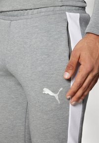 Grå sweatpants med hvid sidepanel, der har et struktureret stof og et lille hvidt Puma-logo på underbenet. Hånd placeret nær bukserne.