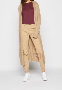 Beigen värinen pörröinen neuletakki, tummanpunainen t-paita, beigen sävyiset kapealahkeiset housut ja valkoiset lenkkarit. Housuissa on vasemman jalan lähellä näkyvä lappu.