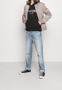 Giacca bomber beige chiaro, felpa nera con cappuccio e logo, jeans azzurri sbiaditi e sneaker nere in tela con suola bianca.