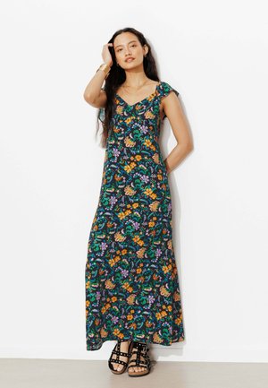 Femme portant une robe maxi fleurie colorée avec de courtes manches volantées et des sandales noires cloutées, debout contre un mur blanc.