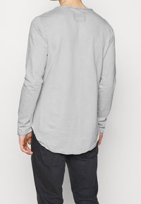 Langärmliges graues T-Shirt aus weicher Baumwolle, mit abgerundetem Saum und einem quadratischen Etikett am hinteren Halsausschnitt. Kombiniert mit dunklen Jeans.