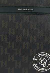 Čierny textúrovaný materiál s opakujúcim sa vzorom loga v tmavo hnedých odtieňoch. Obsahuje striebornú logovú dosku a kruhovú grafickú etiketu.