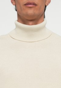Beige coltrui met een geribbelde textuur. Heeft een aansluitend ontwerp en een hoge, gestructureerde kraag. Geen zichtbare patronen.